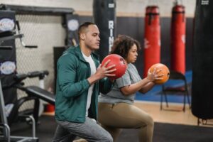 Side view of concentrated young ethnic personal trainer with plus size African American female client doing lunges with medicine balls in modern sport club